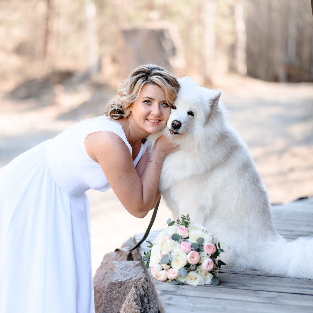 a beautiful bride with curly hair stands beside her white dog. in her hands she holds a wedding